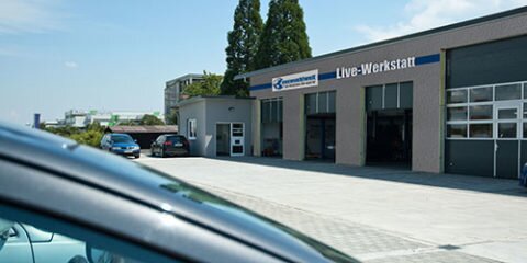 Das Bild zeigt das Gebäude mit der Aufschrift „Live-Werkstatt“ von auswuchtwelt, davor mehrere geparkte Autos auf einem großen Hof.