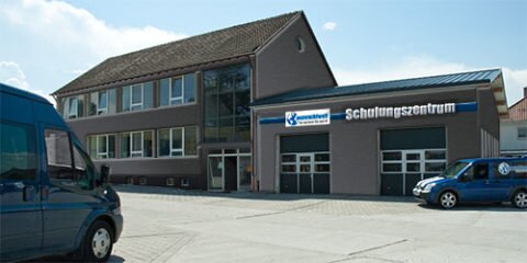 Das Bild zeigt ein Gebäude mit der Aufschrift „Schulungszentrum“ und dem Logo von auswuchtwelt, davor zwei blaue Transporter auf dem Parkplatz.