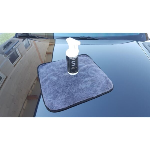 Auf einer glänzenden Motorhaube eines Autos liegt ein graues Mikrofasertuch, auf dem mittig eine weiße Sprühflasche mit dunklem Etikett steht.