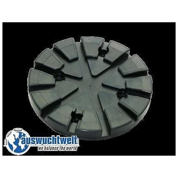 Das Bild zeigt ein rundes Objekt aus schwarzem Material. Es hat eine flache, runde Form mit mehreren erhöhten, trapezförmigen Segmenten, die ringsherum angeordnet sind. Einige dieser Segmente sind leicht gebrochen oder abgesplittert. Das Objekt liegt auf einem weißen Hintergrund. Unten links im Bild ist ein Logo mit der Aufschrift „auswuchtwelt“ und dem Untertitel „we balance the world“ zu sehen. Das Logo zeigt eine stilisierte Weltkugel mit einem Zahnkranz. Das Objekt könnte ein Teil eines technischen Geräts sein, möglicherweise ein Auswuchtgewicht oder ein ähnliches Bauteil.