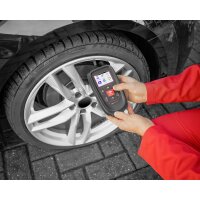 Eine Person in rotem Overall hält ein Diagnosegerät mit Bildschirm und Tasten vor einem Autoreifen. Der Reifen ist schwarz, montiert auf einer hellen Felge mit fünf doppelten Speichen. Das Fahrzeug und der Boden aus Pflastersteinen sind ebenfalls sichtbar. Das Diagnosegerät zeigt verschiedene Symbole auf dem Bildschirm.