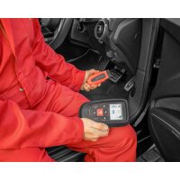 Eine Person trägt einen roten Overall und sitzt auf dem Fahrersitz eines Autos. Sie hält in der linken Hand ein rechteckiges Diagnosegerät mit einem beleuchteten Display, auf dem mehrere Symbole zu sehen sind. In der rechten Hand hält die Person einen USB-Stick oder ein ähnliches Zubehör, das mit dem Diagnosegerät verbunden zu sein scheint. Der Innenraum des Autos zeigt den Bereich unter dem Armaturenbrett mit Gas- und Bremspedal. Die Tür ist geöffnet, und auf dem Lautsprecher im Türbereich steht der Schriftzug „BANG & OLUFSEN“.