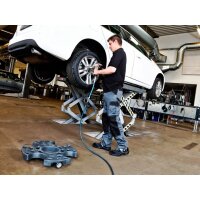In einer Werkstatt arbeitet ein Mann an einem weißen Auto, das auf einer Hebebühne angehoben ist. Der Mann trägt Arbeitskleidung und benutzt ein Werkzeug, um an einem Rad des Autos zu arbeiten. Im Vordergrund steht ein rollbares Werkzeugtablett. Im Hintergrund sind Werkbänke, Werkzeug und Lüftungsrohre zu sehen.