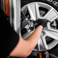 Eine Person mit schwarzem Handschuh befestigt ein Messgerät an einer silbernen Autofelge, der Fokus liegt auf dem Autoreifen und der Hand, im Hintergrund sind Teile von technischem Gerät zu sehen.