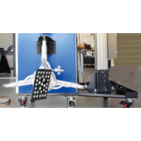 Das Bild zeigt ein technisches Prüfgerät mit Rollen auf einem grauen Boden, eine blaue Metallfläche im Hintergrund und eine Metallvorrichtung mit einem schwarzen Bauteil mit weißen Punkten an der Vorderseite.