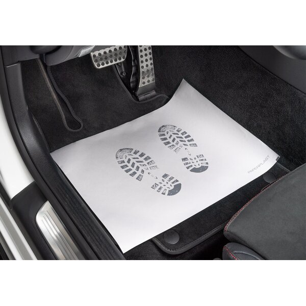 Das Bild zeigt den Fußraum eines Autos auf der Fahrerseite mit den drei Pedalen Kupplung, Bremse und Gas. Auf dem schwarzen Teppichboden liegt eine weiße Papiermatte mit zwei aufgedruckten schwarzen Schuhabdrücken. In der rechten unteren Ecke der Matte steht das Wort „PAPERPLAST“.