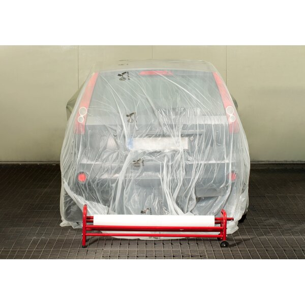Ein Auto steht in einer hellen Garage oder Werkstatt und ist vollständig mit einer dünnen, transparenten Plastikfolie abgedeckt. Vor dem Auto befindet sich ein roter Abroller für die Plastikfolie. Das Auto ist von hinten zu sehen, die Rückleuchten und das Nummernschild sind durch die Folie erkennbar.
